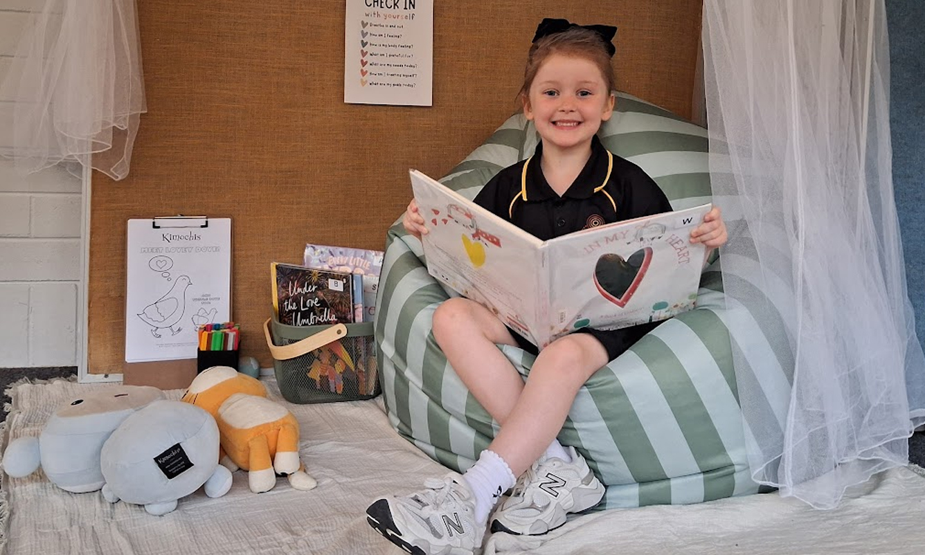 A smiling student stands on a log under a tree. A playground is visible in the background.